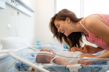 Smiling happy mother looking at baby with love in transparent hospital crib. Mother and baby in...