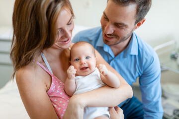 Happy smiling mother and father holding their newborn baby in hospital room. Newborn baby with blue eyes looking at camera.