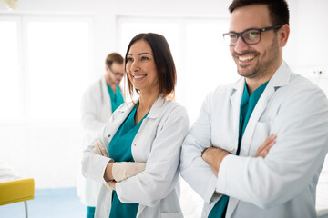Two adult smiling doctors wearing medical scrubs and lab coats standing with arms crossed side by...