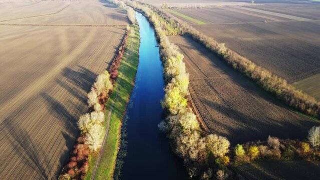 Drone flies above a river surrounded with empty agricultural fields and trees