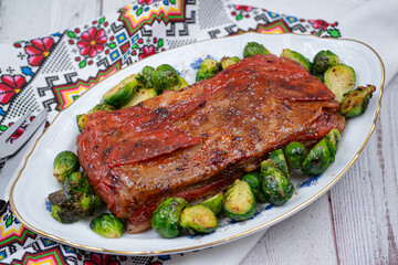Vegan pork belly roast made with seitan and starch, served on oval plate along with roasted Brussels sprouts. Wooden table,  colorful tablecloth