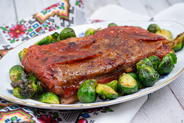 Vegan pork belly roast made with seitan and starch, served on oval plate along with roasted Brussels sprouts. Wooden table,  colorful tablecloth