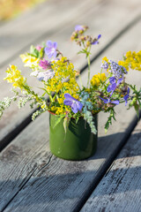 Bouquet of bright colourful summer wildflowers in a green metal mug on wooden background. Sunny morning. Baikal lake, Siberia