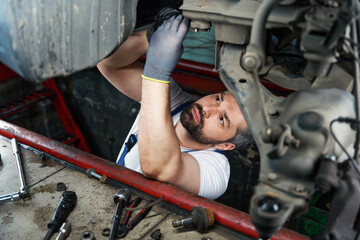 Focused automotive service technician is fixing automobile in inspection pit - obrazy, fototapety, plakaty