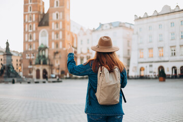 Attractive female tourist is exploring new city. Redhead woman pointing finger on Market Square in Krakow. Traveling Europe in autumn. St. Marys Basilica. Active lifestyle