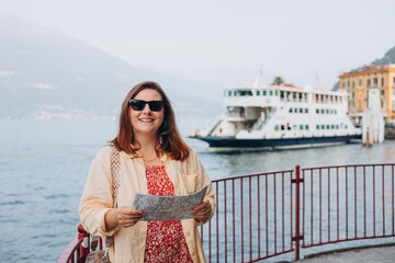 Happy brunette 30s woman enjoying summer holidays. Concept of vacation. Varenna, Italy. Lake Como. Attractive female tourist is exploring new city. Redhead girl holding a paper map