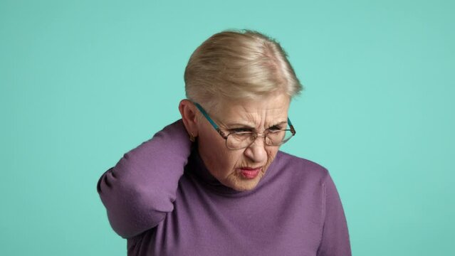 Old woman with light make-up in purple turtleneck wincing in pain over blue background. Age-related health problems, elderly lady suffering from neck pain. High quality 4k footage