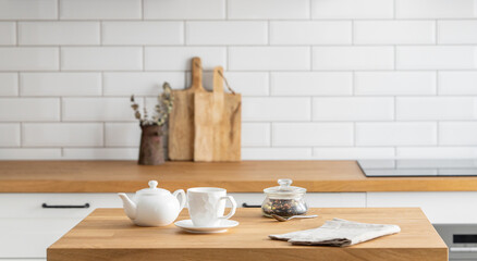 Wooden oak table with a cup of tea and a kettle  in front of the kitchen with a white brick background. - obrazy, fototapety, plakaty