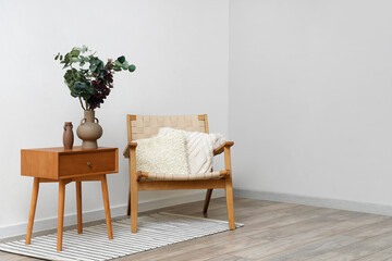 Vase with green eucalyptus branches on end table and comfortable armchair near white wall