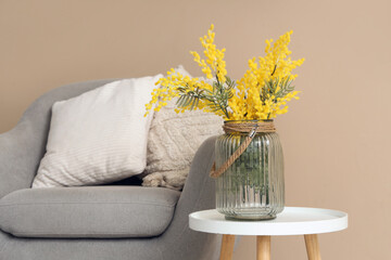 Vase with beautiful mimosa flowers on end table and grey armchair near beige wall