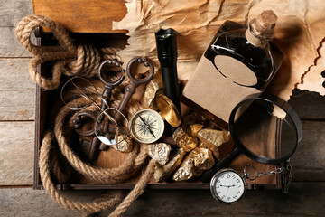 Box with travel equipment, golden nuggets and bottle of rum on brown wooden background - obrazy, fototapety, plakaty