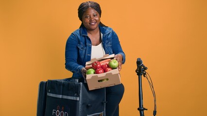 Female african american courier gently transporting fruit basket from food delivery bag. Excited...