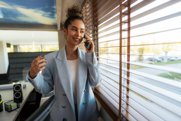 In a corporate setting, a female executive with curly hair sips coffee, looks out the office window, and discusses business matters on the phone.