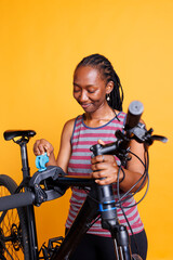 Black female cyclist inspecting her bicycle broken frame while placed on repair stand against isolated backdrop. Young woman preparing to use specialized tools for adjustments and repairs.