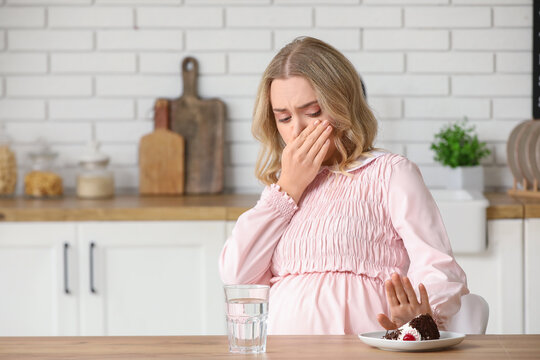 Young pregnant woman with piece of cake suffering from toxicosis at table in kitchen