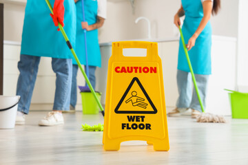 Team of young janitors mopping floor with caution sign in kitchen, closeup