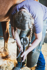 Quarter horse getting hoof shaped by a female farrier with two pigtail braids in a dusty stall in...