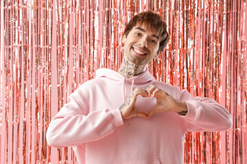 Happy young man making heart with hands and tinsel curtain on pink background. Valentine's day...