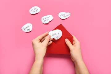 Female hands with red envelope and declarations of love on pink background, closeup. Valentine's Day celebration