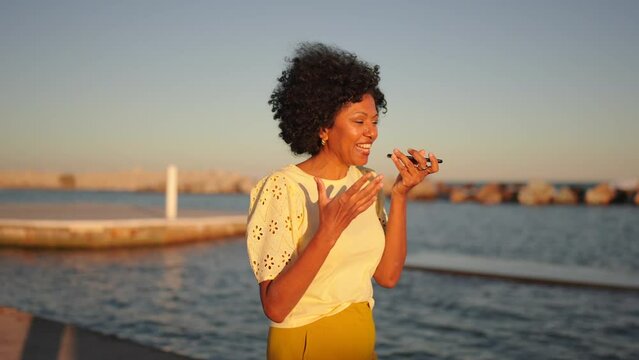 Video of an african woman smiling while recording a voice message with the mobile next to the sea during sunset