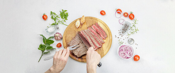 Fototapeta na wymiar Woman cuts grilled steak. Female hands with knife and two pronged fork. Fresh herbs, vegetables