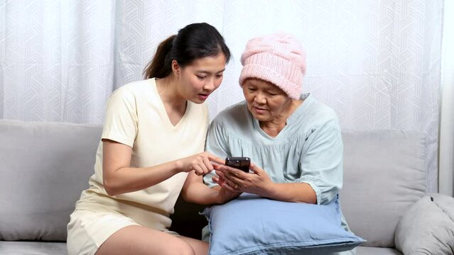 Asian caregiver nurse female taking care of cancer chemotherapy elderly senior woman at home, young daughter and sickness retired mother happy learning mobile phone application free time in holiday