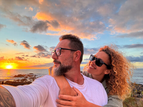 Happy and romantic couple of traveler take selfie picture with modern smartphone at the beach with amazing orange sunset in background. People and summer travel holiday vacation. Happy lifestyle