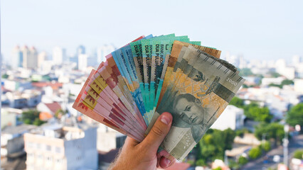 Man's Hand holding rupiah money against the backdrop of the capital of Indonesia. - obrazy, fototapety, plakaty