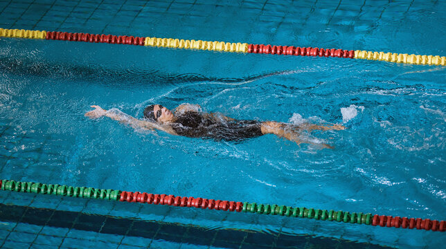 Female athlete in action, performing the backstroke swim technique in the indoor lap pool. Competitive back crawl stroke concept. - Powered by Adobe