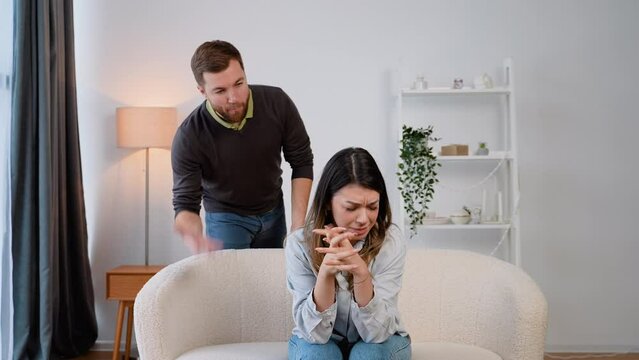 Hopeless young woman sitting on the sofa at home, while her husband abuser screaming at her, expressing aggression. Domestic violence and emotional abuse concept