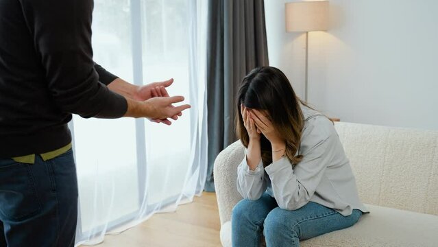 Upset wife sit on couch crying listening to furious husband yelling, unhappy couple have fight or disagreement at home