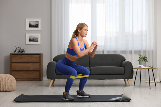Athletic woman doing squats with fitness elastic band near laptop on mat at home