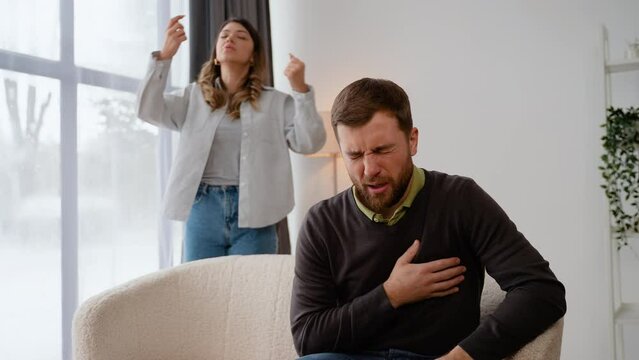 Aggressive woman screaming at scared husband at home. A man holds on his chest, feeling heart pain. Heart attack, myocardial infarction