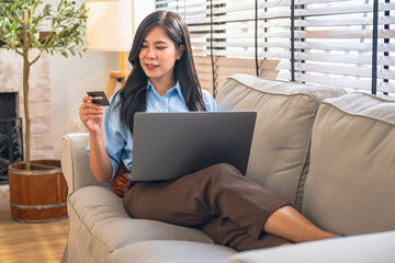 happily relaxed young woman sits on a sofa using computer and debit card shopping on line