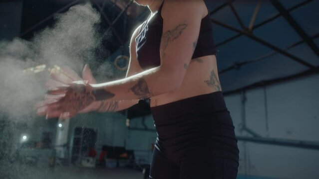 Young female weightlifter with tattooed body applying chalk powder to hands before training in a gym, rubbing palms together and making dust clouds flying in the air. Tilt-up shot in slow mo