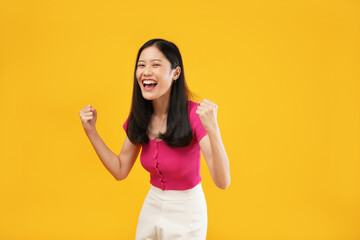 Portrait of a young Asian woman casually dressed woman screaming yes and clenching her fists, winner's gesture concept. Isolated on a yellow background.  - obrazy, fototapety, plakaty
