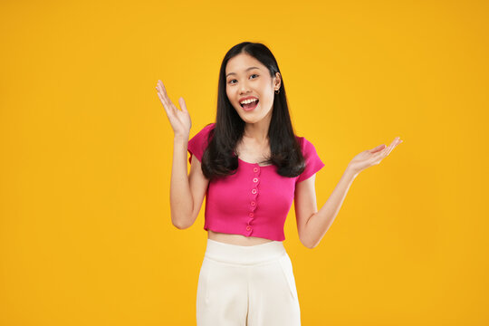 Photo of excited and surprised young asian woman saying wow with amazement, raising hands up near face, standing against yellow background