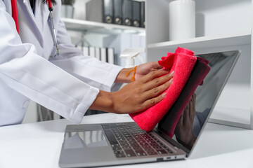 medical professional, likely a female doctor, sanitizing a desk with a red cloth and disinfectant spray, emphasizing cleanliness in the medical environment.