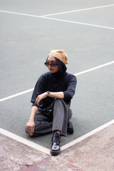 Asian female model in jeans and a black shirt, sitting on a tennis court with a bandana and glasses