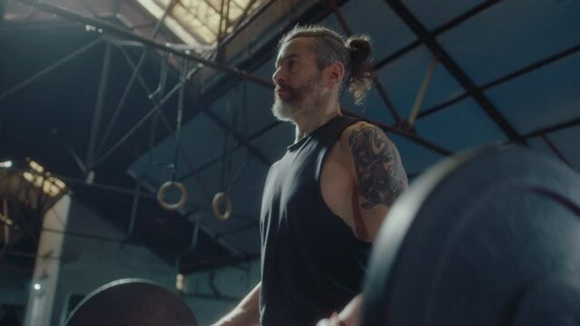 Muscular mature man with tattooed arms doing barbell bicep curls during weightlifting training session in a gym. Zoom shot