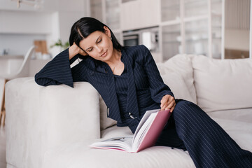 Contemplative woman in pinstripe suit lounging on a sofa while engrossed in reading a book
