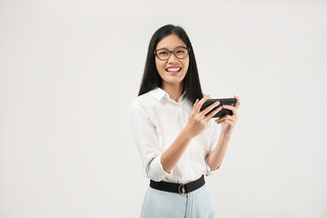 This portrait of a young Asian woman wearing a white shirt and glasses, with a positive expression playing video games on a modern smartphone, isolated on a white background. Modern technology concept