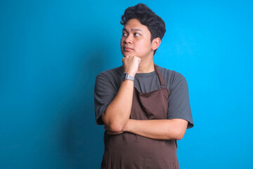 Asian barista man in brown apron touching his chin and looking to the side over blue background