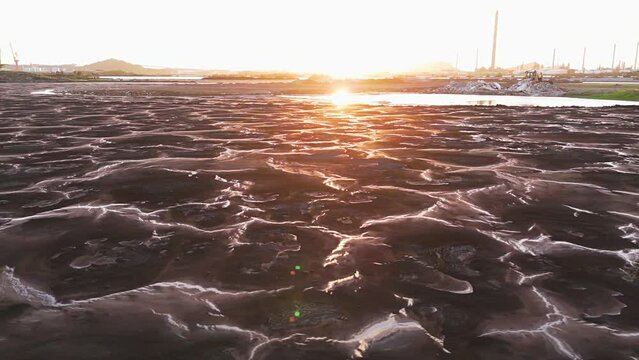 Sunlight glistens against polluted water in asphalt lake at golden hour with oil refinery behind barren ground