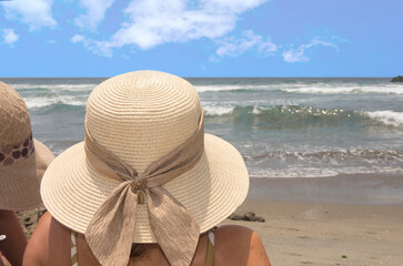 Woman in straw hat sitting on the shore looking at the sea under midday sky