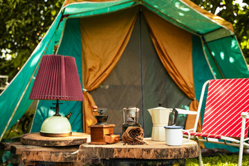 selective focus of vintage coffee set on the old wooden table In front of the retro cabin tent, Antique oil lamp, retro chairs, Group of camping tents, and soft focus.