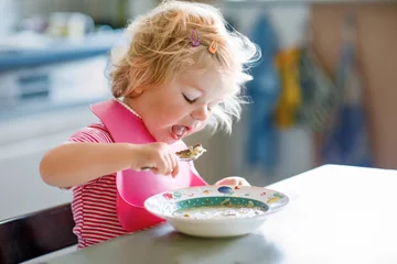 Schilderijen op glas Adorable baby girl eating from spoon vegetable noodle soup. Healthy food, child, feeding and development concept. Cute toddler child with spoon sitting in highchair and learning to eat by itself © Irina Schmidt