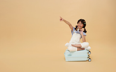 asian child sitting on suitcase pointing finger at copy space and look through binoculars isolated...