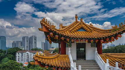 Obraz premium A Chinese Thean Hou Temple against a blue sky, clouds, city skyscrapers. The elegantly curved roof is decorated along the edge with row of red traditional lanterns. Stairs with white carved handrails
