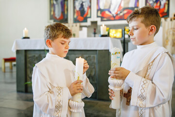 Two little kids boys receiving his first holy communion. Happy children holding Christening candle. Tradition in catholic curch. Kids in a church near altar. Siblings, brothers in white gowns.